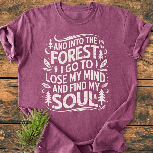 And Into The Forest I Go - Vintage Wash Tee, featuring white graphic text with nature-themed illustrations on maroon fabric, displayed flat with a pine branch accent on a wooden surface.