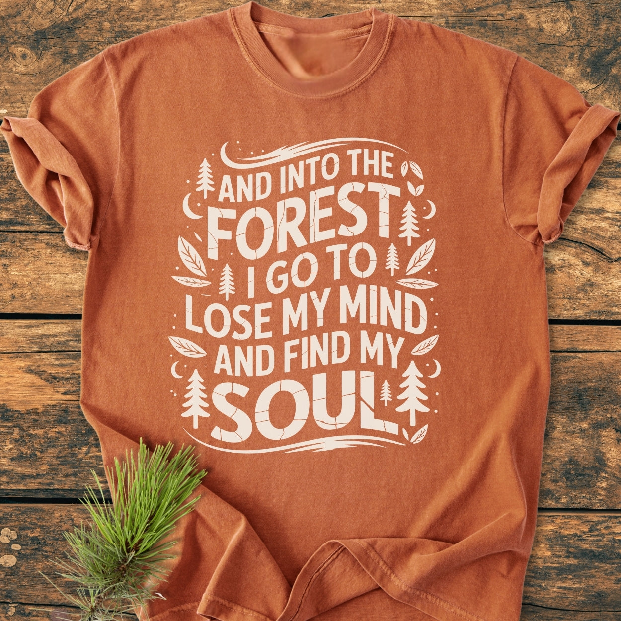 And Into The Forest I Go - Vintage Wash Tee featuring an outdoor-inspired white graphic with nature motifs, laid on wood with a pine sprig, showcasing its relaxed fit and durable stitching.