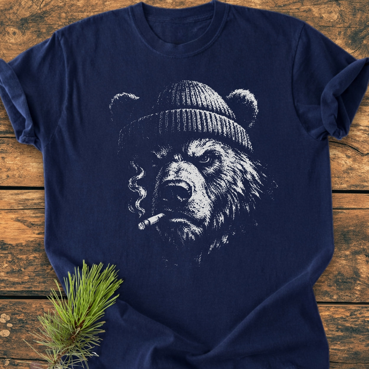 Smoking Bear - Vintage Wash Tee: Navy-blue T-shirt featuring a sketched grizzly bear with a beanie and cigarette. Laid on wood with a spruce twig beside. Made from soft-washed, pre-shrunk ring-spun cotton.