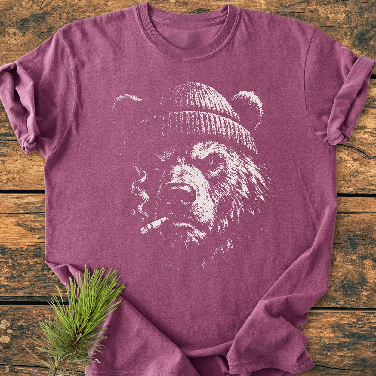 Smoking Bear - Vintage Wash Tee featuring a grizzly bear in a beanie with a cigarette, printed on a heather-purple T-shirt, laid on a wooden background with a pine branch accent.
