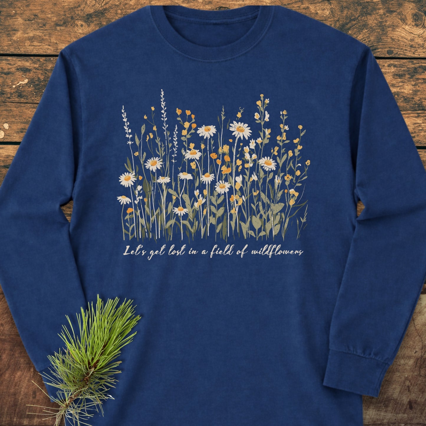 Let's Get Lost In A Field Of Wildflowers Long-Sleeve Tee features a navy-blue design with a meadow-style wildflower print and phrase, laid flat on a wooden surface with a pine sprig beside it.