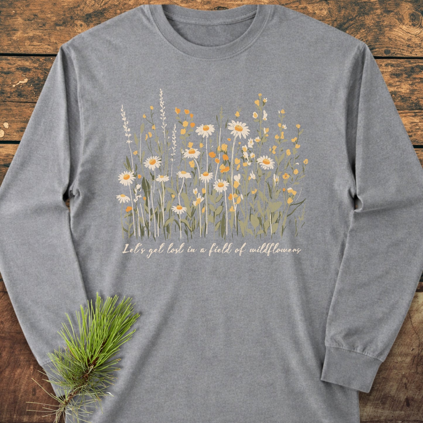 Let's Get Lost In A Field Of Wildflowers Long-Sleeve Tee, heather-gray, featuring a white and yellow wildflower print with cursive slogan, styled on wood with a pine branch accent.