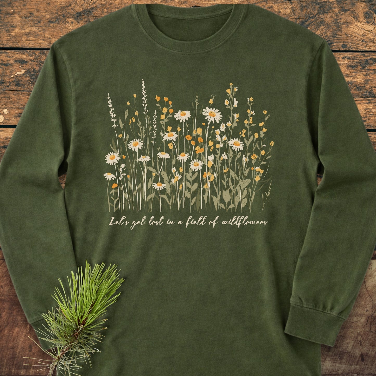Let's Get Lost In A Field Of Wildflowers Long-Sleeve Tee, showcasing wildflower illustrations and a slogan on forest-green cotton, laid on wood with a pine twig accent for a rustic touch.