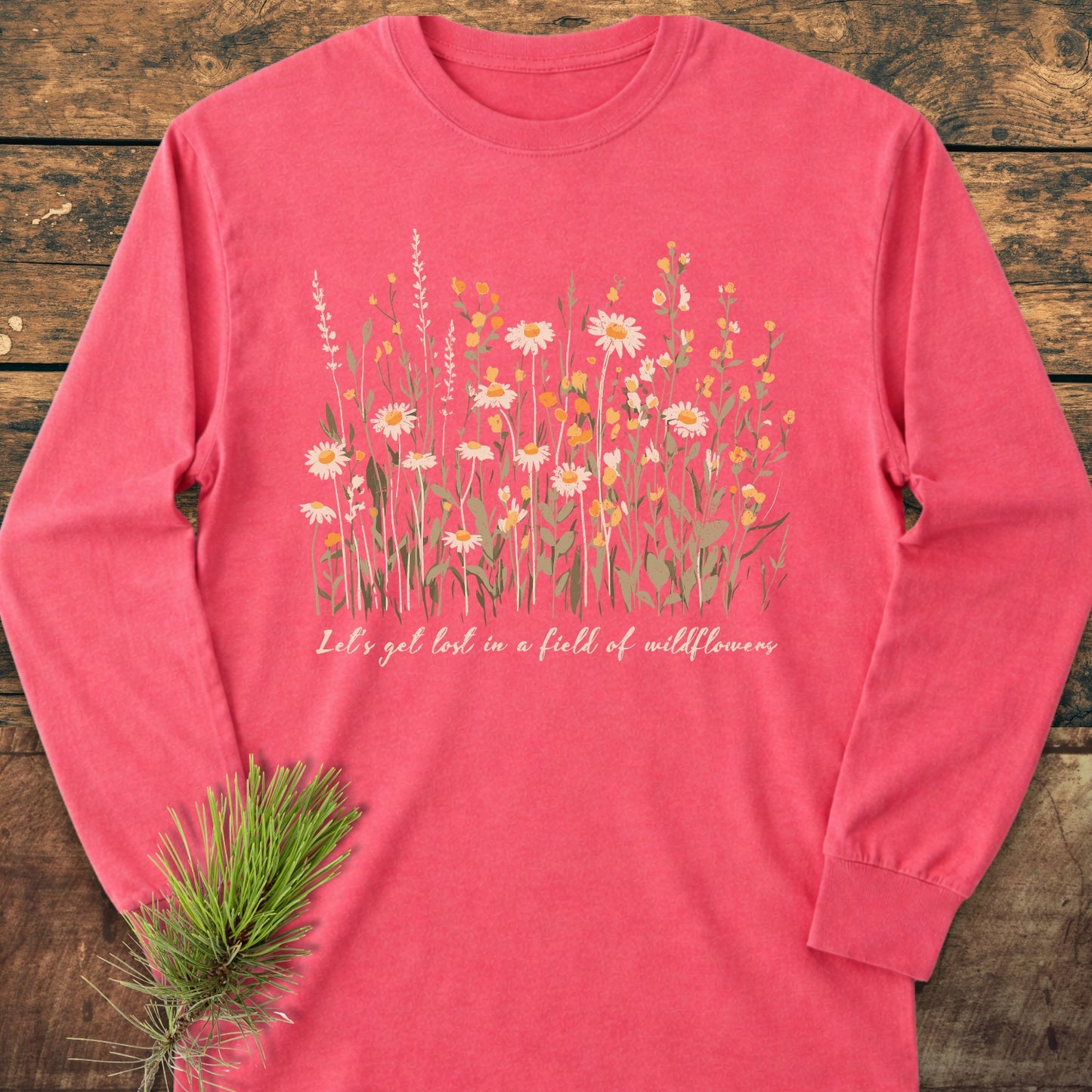 Let's Get Lost In A Field Of Wildflowers Long-Sleeve Tee styled on rustic wood, featuring vintage-washed fabric with wildflower illustrations and text, accompanied by a pine sprig.