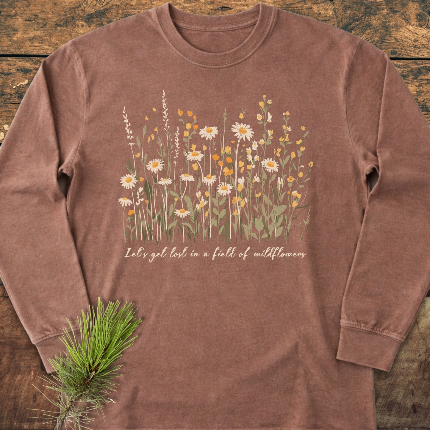 Let's Get Lost In A Field Of Wildflowers Long-Sleeve Tee, featuring hand-drawn daisies and blooms with script text on a rustic wooden surface, alongside a small pine sprig.