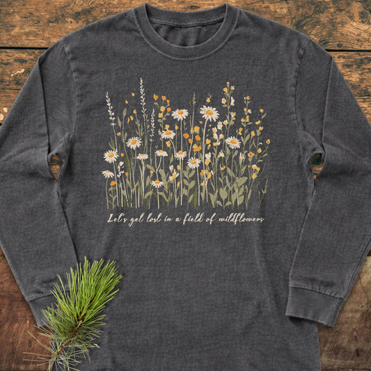 Let's Get Lost In A Field Of Wildflowers Long-Sleeve Tee features a wildflower illustration and slogan on a charcoal-gray shirt, laid flat with a pine sprig beside it.