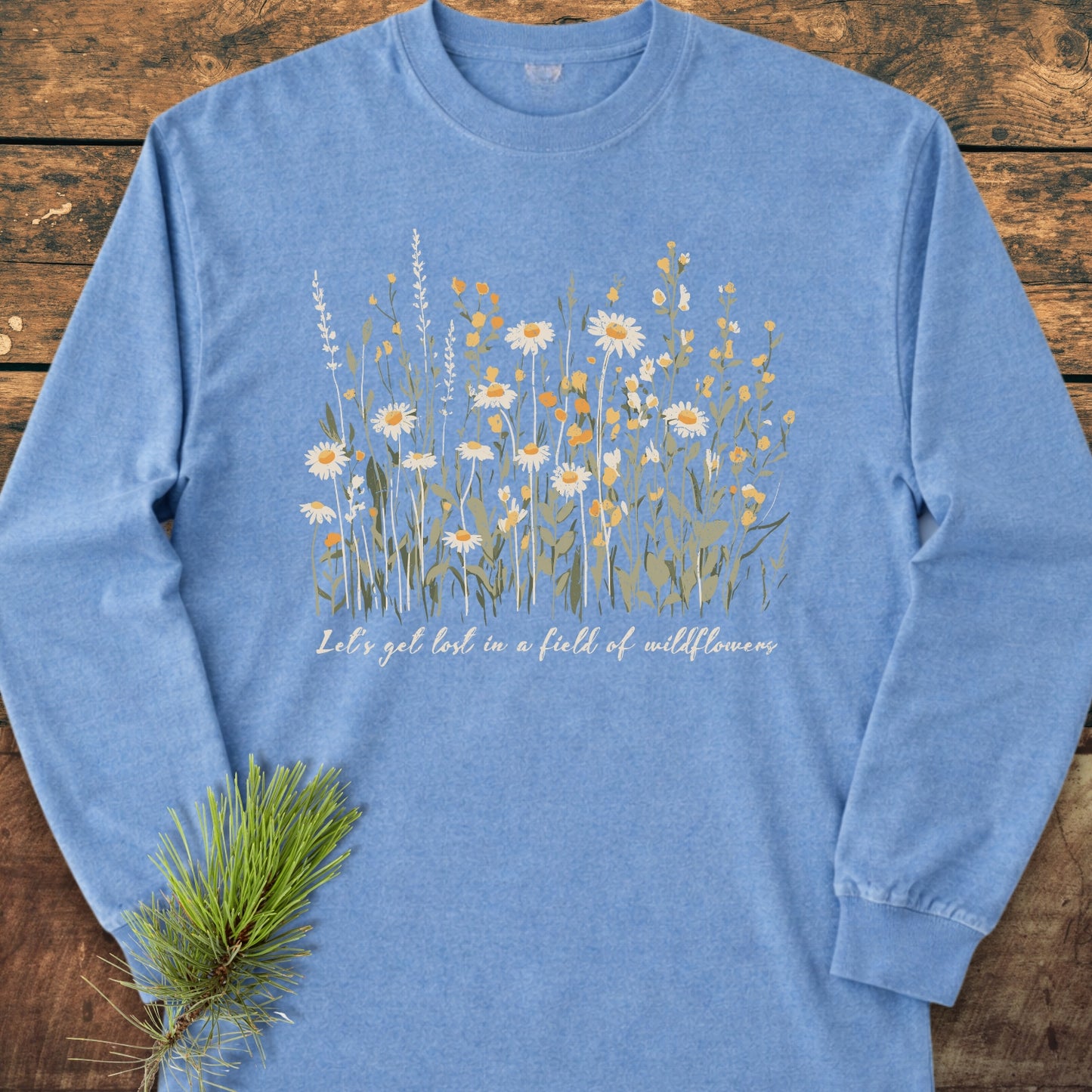 Let's Get Lost In A Field Of Wildflowers Long-Sleeve Tee featuring stylized wildflowers and a cursive slogan, laid flat on wood with a pine branch accentuating its rustic charm.