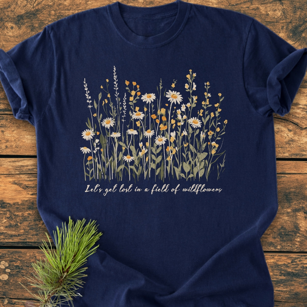 Let's Get Lost In A Field Of Wildflowers - Vintage Wash Tee featuring hand-drawn wildflowers and slogan, arranged on wooden planks with pine sprig, showcasing its soft, garment-dyed, ring-spun cotton fabric.