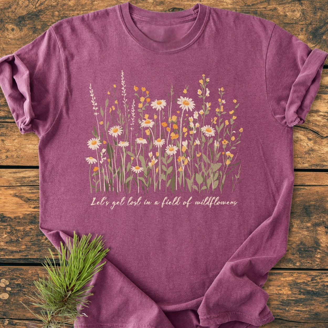 Let's Get Lost In A Field Of Wildflowers - Vintage Wash Tee, featuring hand-drawn wildflowers and phrase, styled with a pine sprig on a wooden surface, highlighting its cozy, relaxed fit.