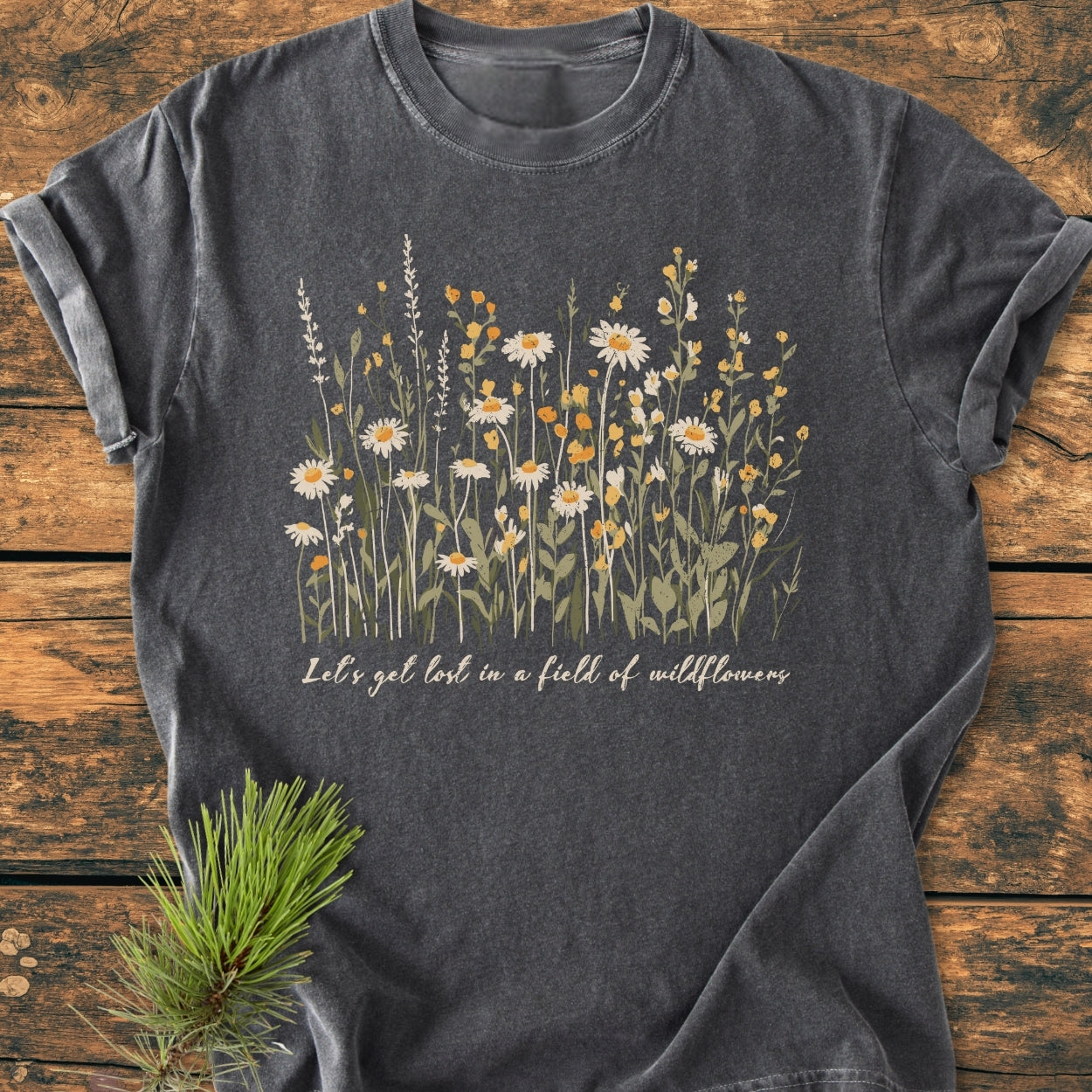Let's Get Lost In A Field Of Wildflowers - Vintage Wash Tee features a line-drawing bouquet of daisies and blossoms with cursive text, laid on a wooden surface, embodying comfort and personalized style.