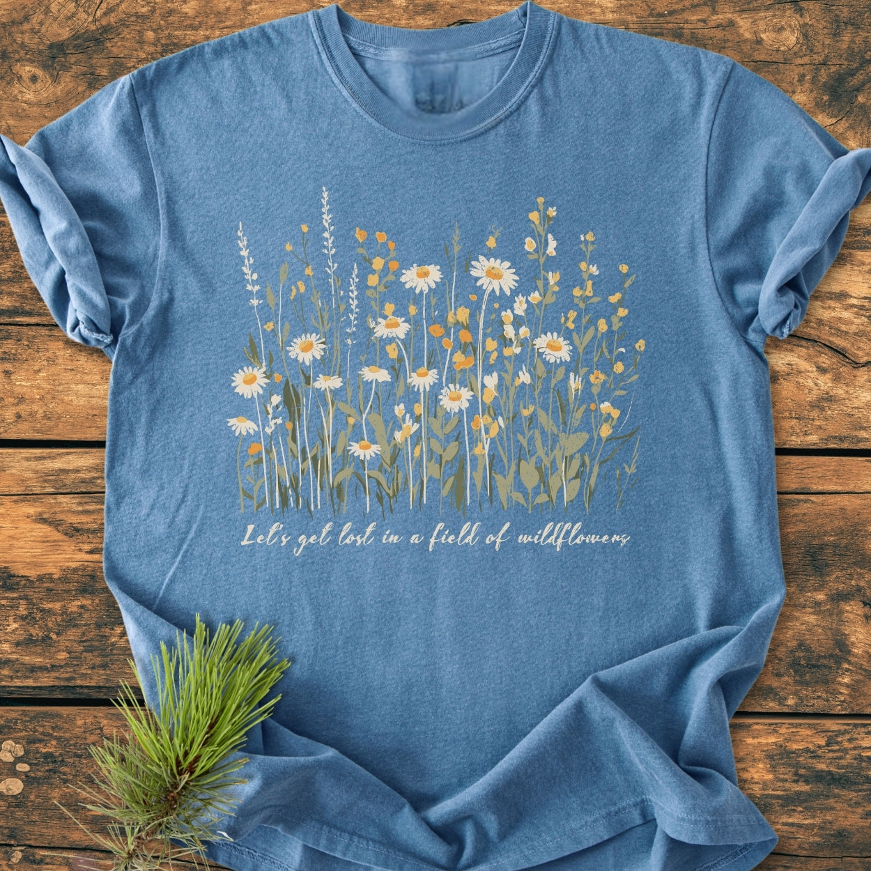 Let's Get Lost In A Field Of Wildflowers - Vintage Wash Tee, featuring soft heather-blue cotton with wildflower design and cursive slogan, laid flat with a rustic pine sprig accent.