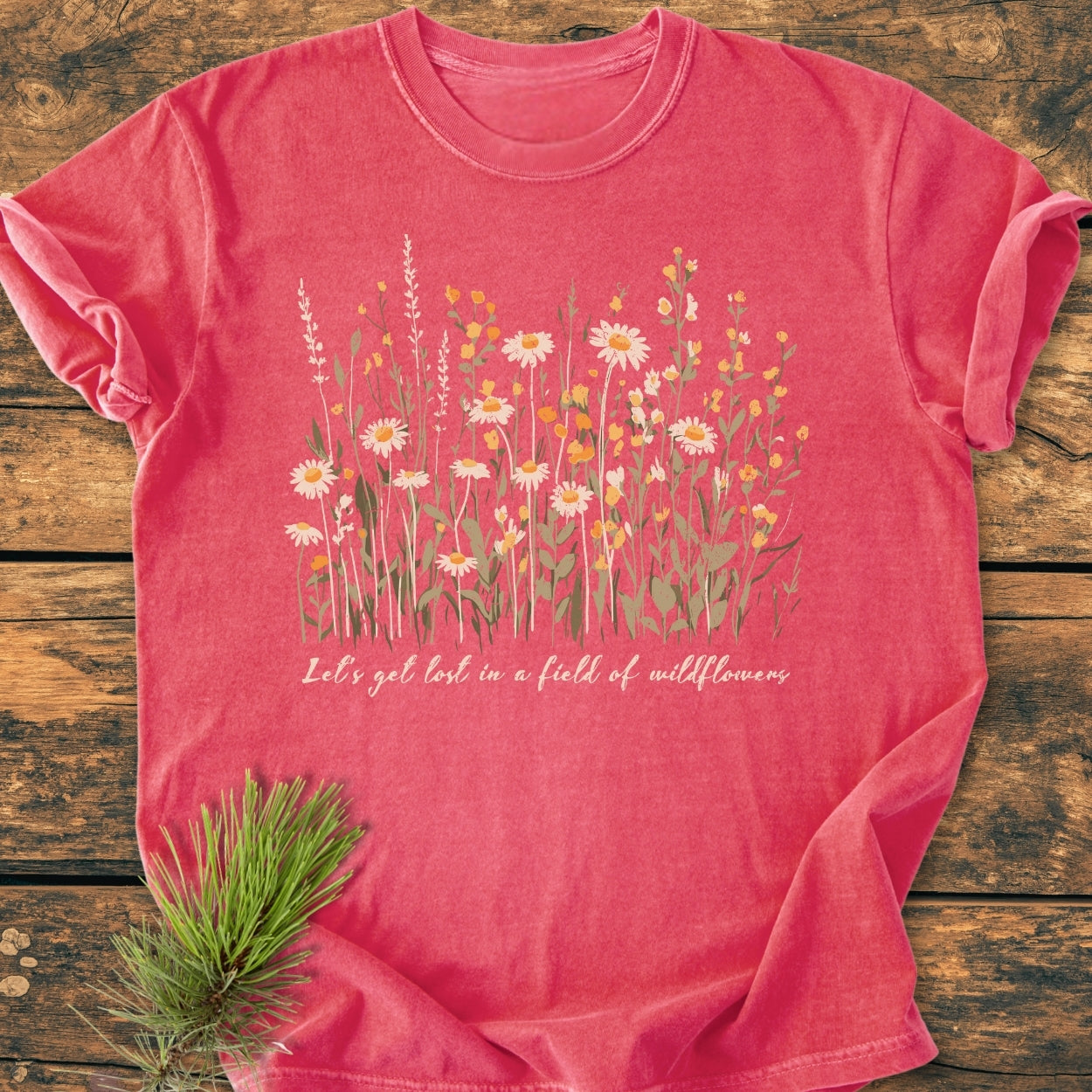Let's Get Lost In A Field Of Wildflowers - Vintage Wash Tee features delicate wildflowers and slogan on a heather-red crew-neck. Laid on wood with a pine sprig accenting the scene.