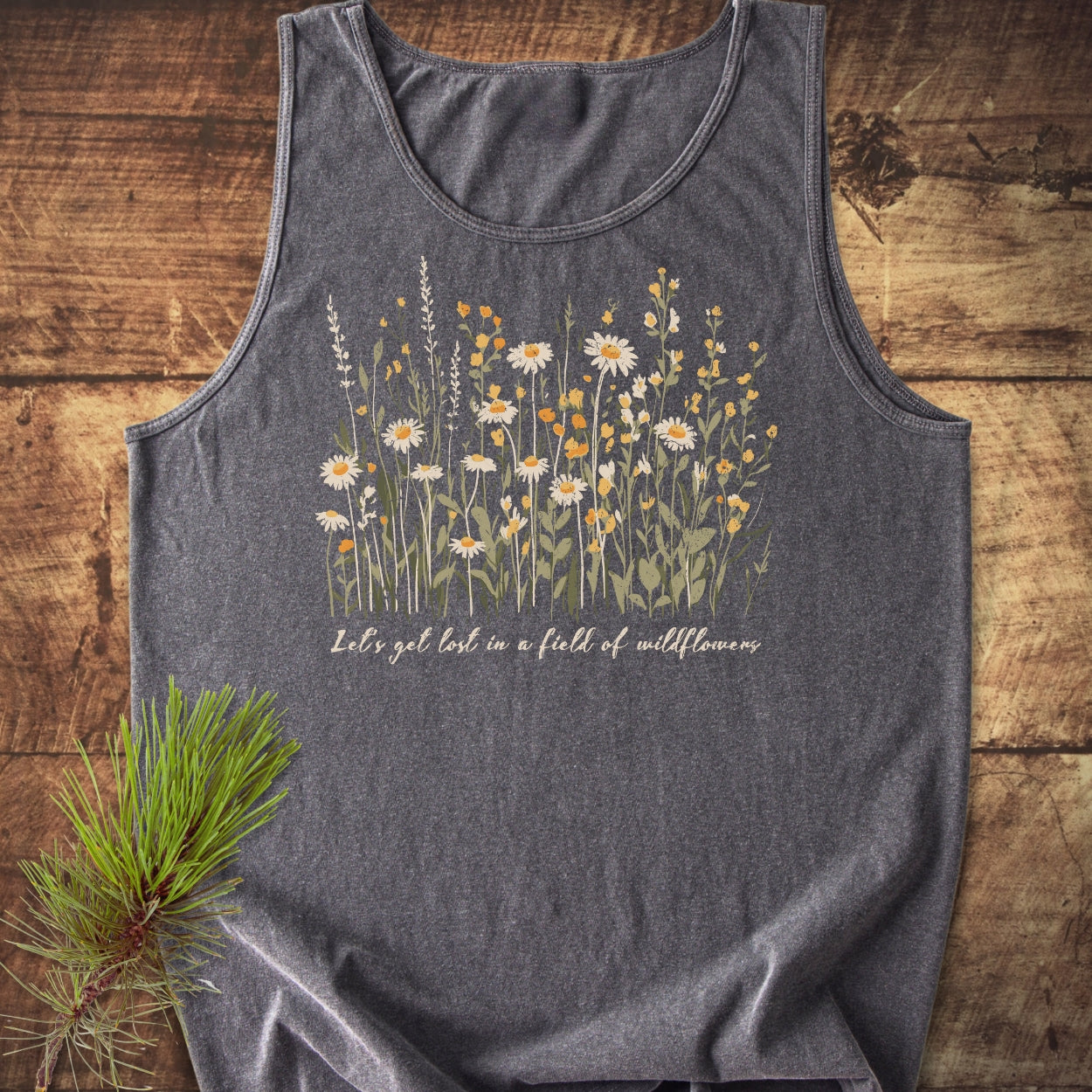 Let's Get Lost In A Field Of Wildflowers Tank Top featuring a daisy and wildflower illustration on charcoal-grey, styled with a pine branch on rustic wood, highlighting comfort and sustainability.
