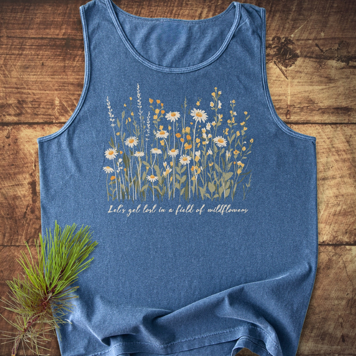 Let's Get Lost In A Field Of Wildflowers Tank Top features a meadow-style floral design and scripted text on a heather-blue sleeveless top, laid flat with a pine branch accentuating its rustic appeal.