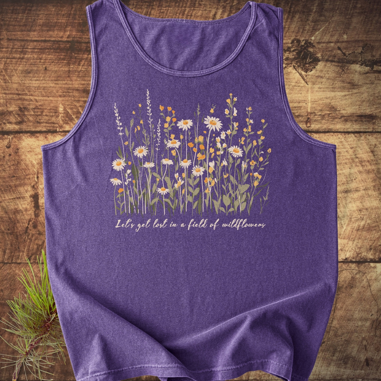 Let's Get Lost In A Field Of Wildflowers Tank Top features a meadow of wildflowers and the phrase on a purple sleeveless design, styled with a pine sprig on a wooden surface.
