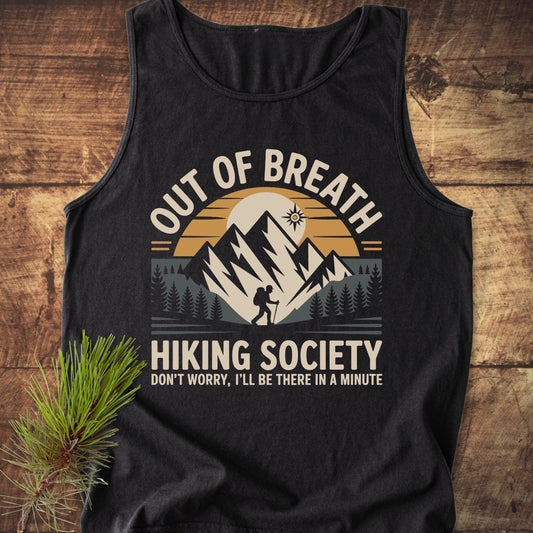 Out Of Breath Hiking Society Tank Top displayed on a wooden surface, features a hiker graphic with mountains, sun, and pine trees. Includes humorous text and a pine sprig beside the shirt.