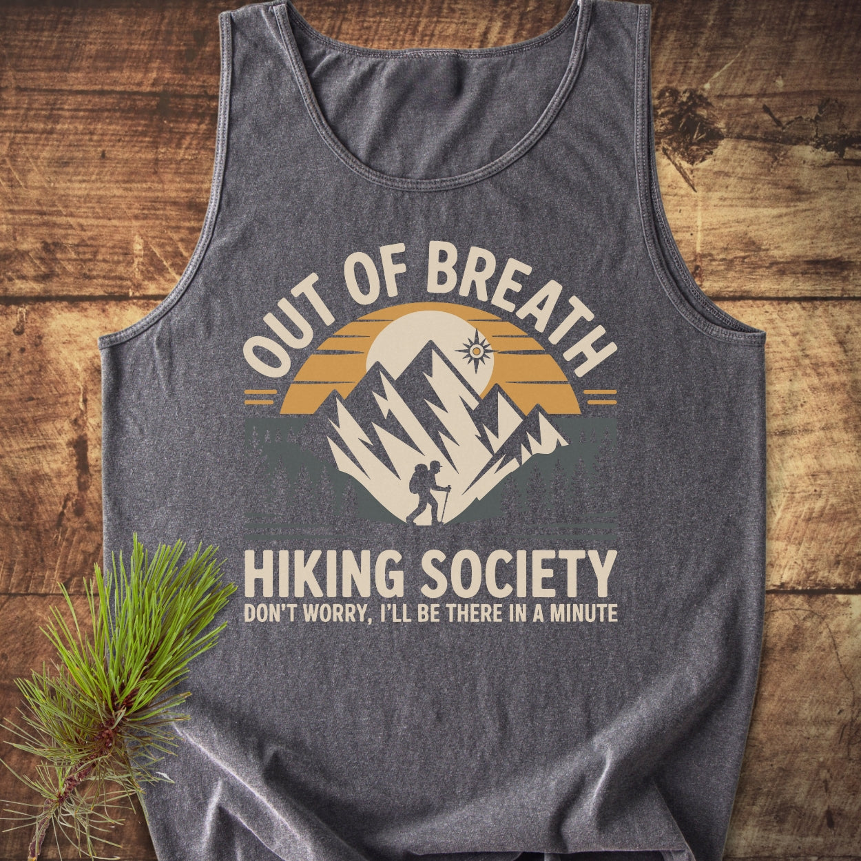 Heather-gray Out Of Breath Hiking Society tank top with mountain-and-sun graphic, featuring a lone hiker. Laid on wood, accompanied by a pine sprig, emphasizing comfort, style, and sustainability.