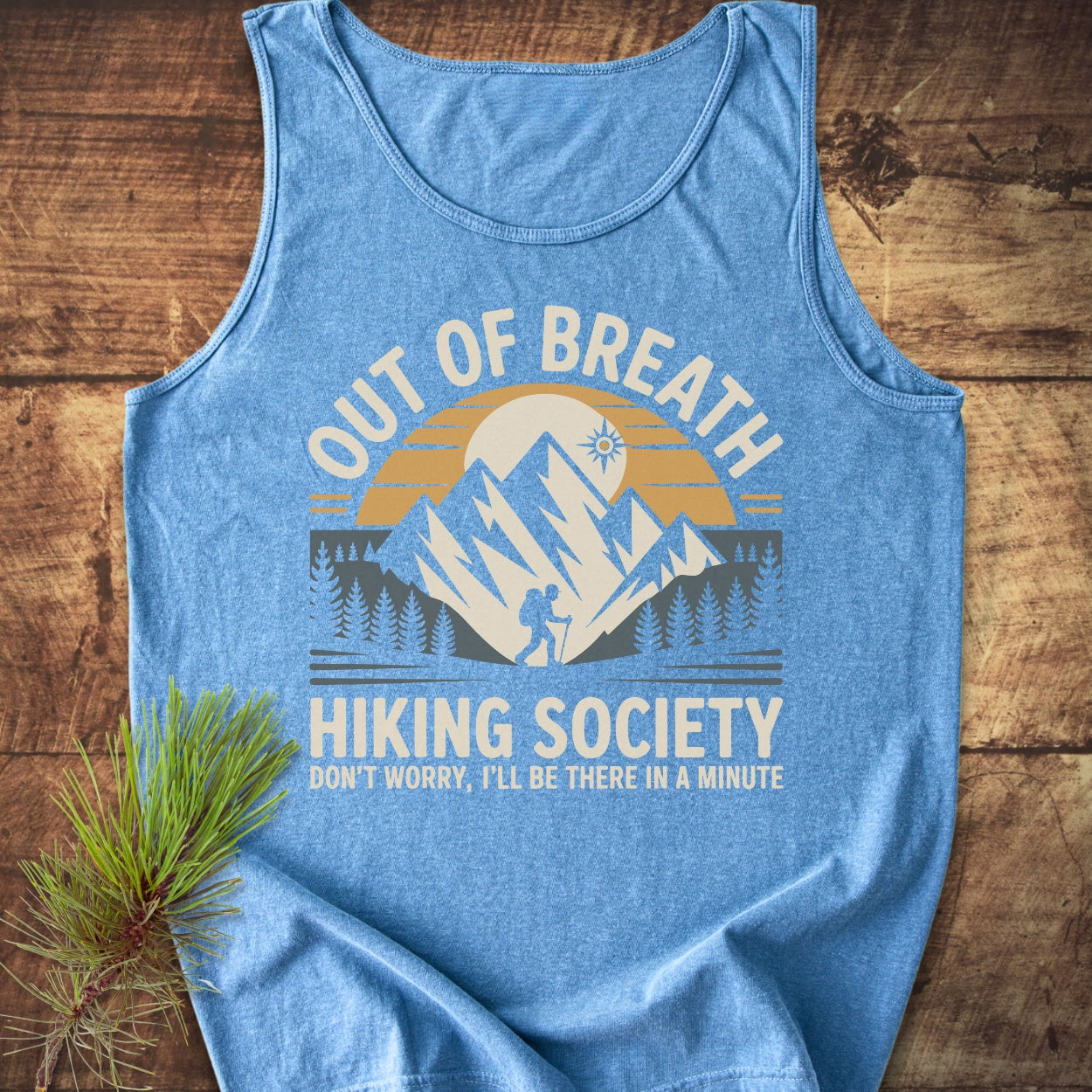 Out Of Breath Hiking Society Tank Top featuring a mountain scene and slogan, laid on wood with a pine sprig. Sleeveless, relaxed fit, showcasing comfort and sustainability with ethical, eco-friendly production.