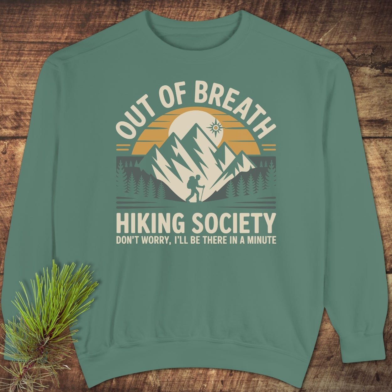 Out Of Breath Hiking Society Sweatshirt featuring a sunrise over mountains, a hiker among pines, and humorous text. Laid on wood with pine sprig, this sweatshirt offers luxurious comfort and style.