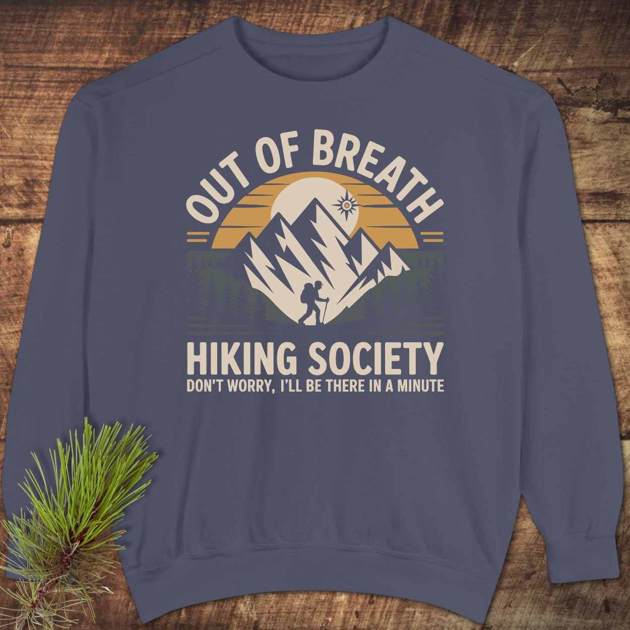 Out Of Breath Hiking Society Sweatshirt on wood, featuring a vibrant outdoors graphic with a hiker and mountains. Includes text DON’T WORRY, I’LL BE THERE IN A MINUTE and a pine branch accent.