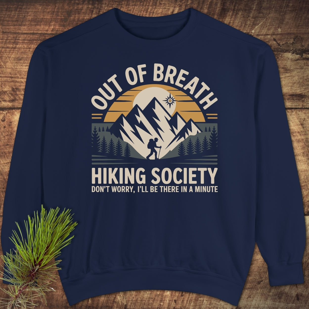Out Of Breath Hiking Society Sweatshirt featuring a stylized mountain scene with a lone hiker, printed on a navy-blue crewneck. Laid flat on wood, accompanied by a pine sprig.
