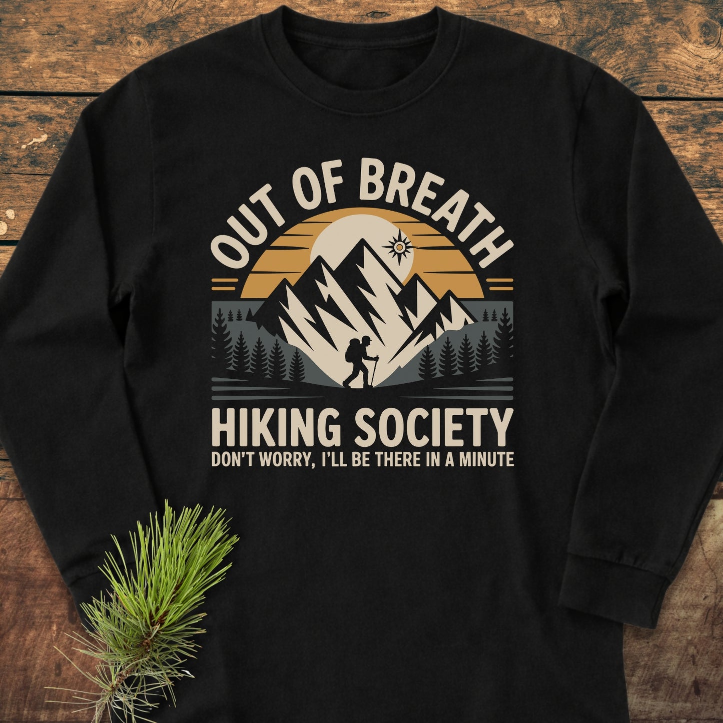 Out Of Breath Hiking Society Long-Sleeve Tee on wooden surface, featuring a stylized outdoor scene with mountains, trees, sun, and hiker, paired with humorous hiking slogan. Made from soft, garment-dyed ring-spun cotton.