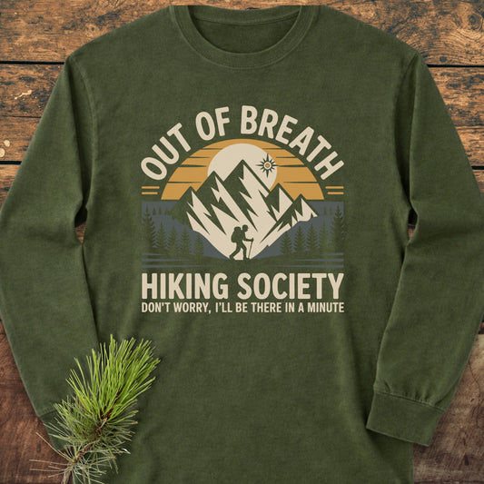 Out Of Breath Hiking Society Long-Sleeve Tee with hiking graphic, ring-spun cotton for comfort, laid on wood, pine branch accentuating its outdoor theme.