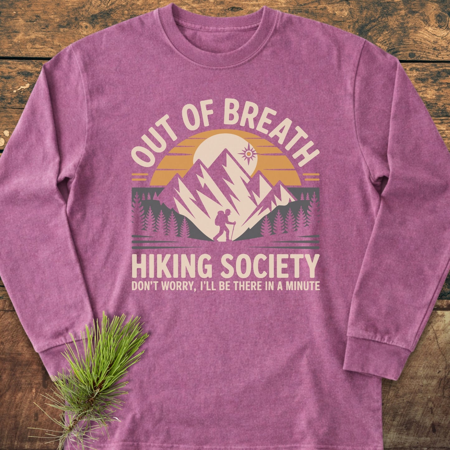 Out Of Breath Hiking Society Long-Sleeve Tee featuring a retro hiking graphic with mountains, trees, and sun. Displayed flat with a pine sprig on rustic wood, highlighting its soft, relaxed fit.