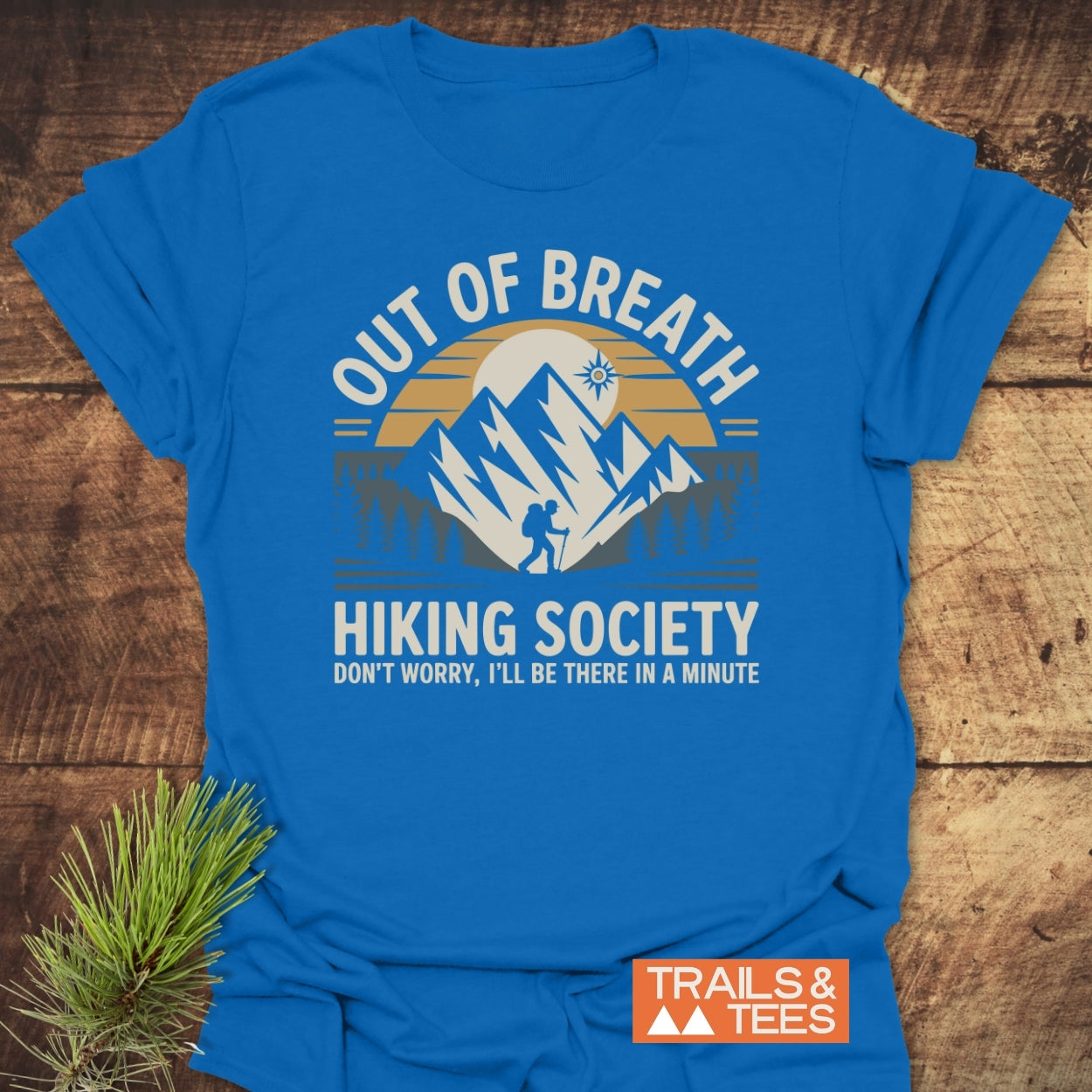Out Of Breath Hiking Society T-Shirt: Blue tee with a mountain scene and humorous text. Accompanied by a pine sprig, highlighting a love for adventure and nature.