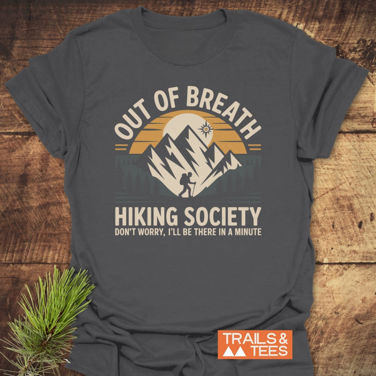 Out Of Breath Hiking Society T-Shirt featuring a sunrise over mountains, hiker silhouette, and playful text. Crafted from soft cotton, it promises comfort for outdoor enthusiasts. Includes TRAILS & TEES brand mark.