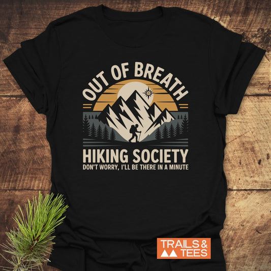 Out Of Breath Hiking Society T-Shirt features a graphic of mountains, pine trees, and a hiker, with playful text. Laid on wood, it’s crafted from soft, durable cotton for outdoor enthusiasts.