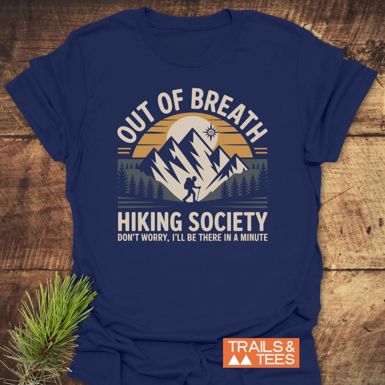 Out Of Breath Hiking Society T-Shirt featuring a backpacker silhouette against jagged mountains and sun, with humorous text, laid flat showcasing its outdoor-themed design and quality craftsmanship.