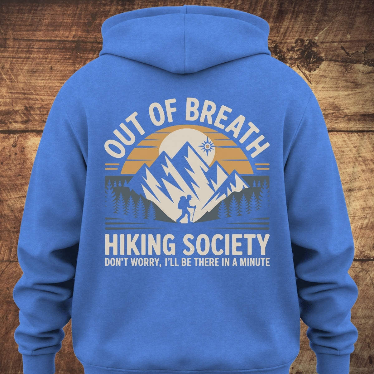 Out Of Breath Hiking Society BACKPRINT on a royal-blue hoodie featuring a sun setting behind mountains with a hiking silhouette and pine trees, complemented by hiking-themed slogans.