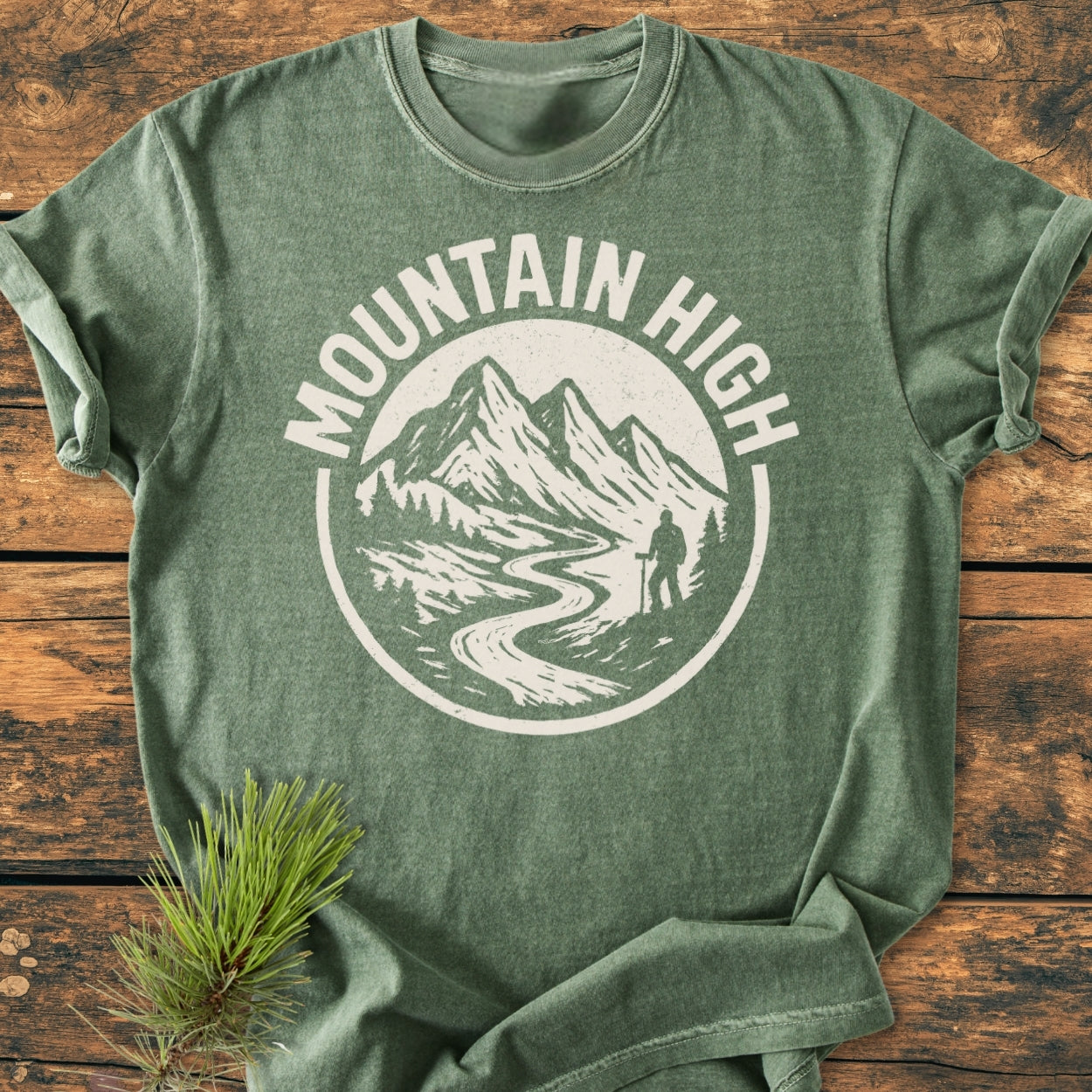 Mountain High - Vintage Wash Tee featuring a white emblem of mountains, pine trees, and a hiker, laid flat on a wooden surface with a pine sprig, highlighting its outdoor-inspired design.