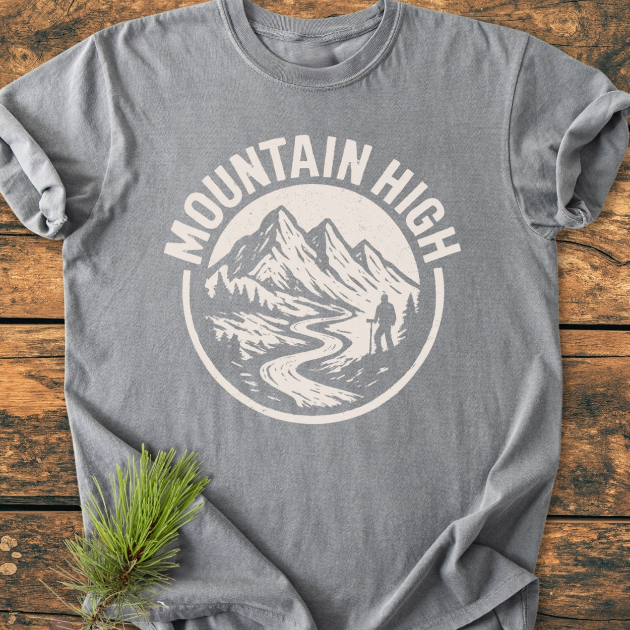 Mountain High - Vintage Wash Tee featuring a circular mountain landscape graphic with peaks, river, trees, and hiker. Laid flat on wood, showcasing its relaxed fit and soft, durable fabric.