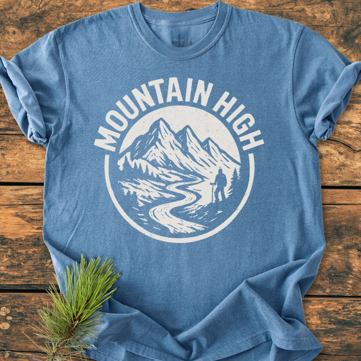 Mountain High - Vintage Wash Tee features a heather-blue color, with a white Mountain High logo depicting peaks, a trail, and hiker, laid flat on wood, paired with a pine sprig.