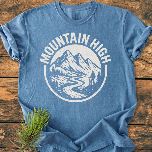 Mountain High - Vintage Wash Tee features a heather-blue color, with a white Mountain High logo depicting peaks, a trail, and hiker, laid flat on wood, paired with a pine sprig.
