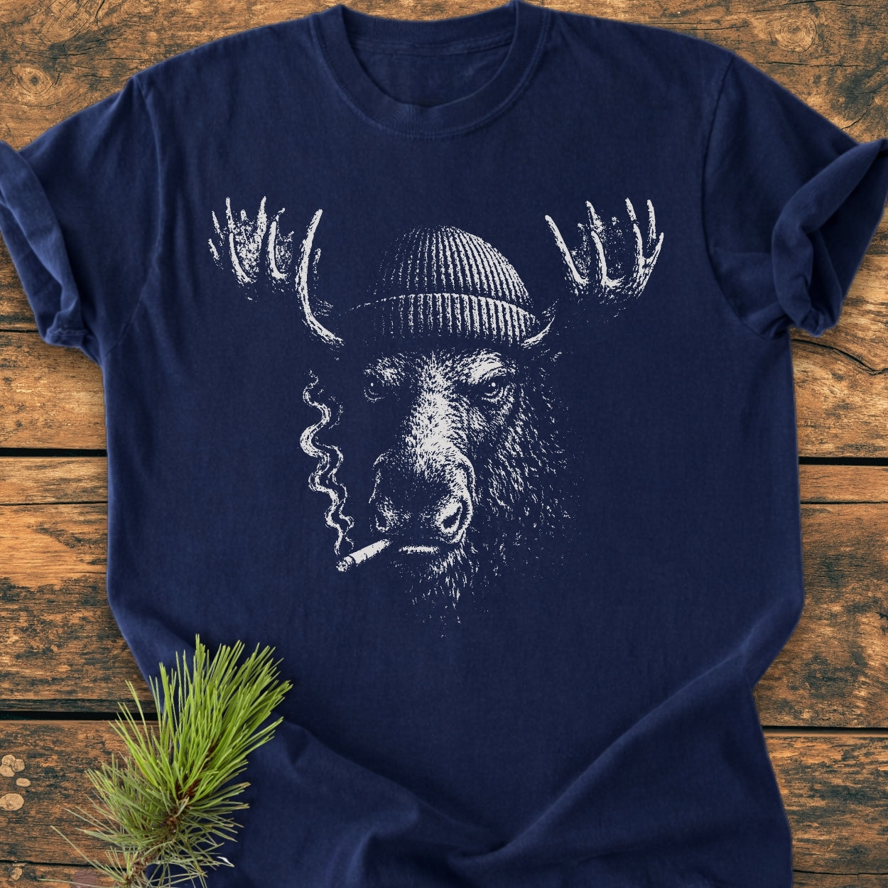 The Slow Burn Moose - Vintage Wash Tee displays a moose in a beanie with a cigarette, printed on a navy-blue shirt, laid on wood with a pine sprig accenting it.