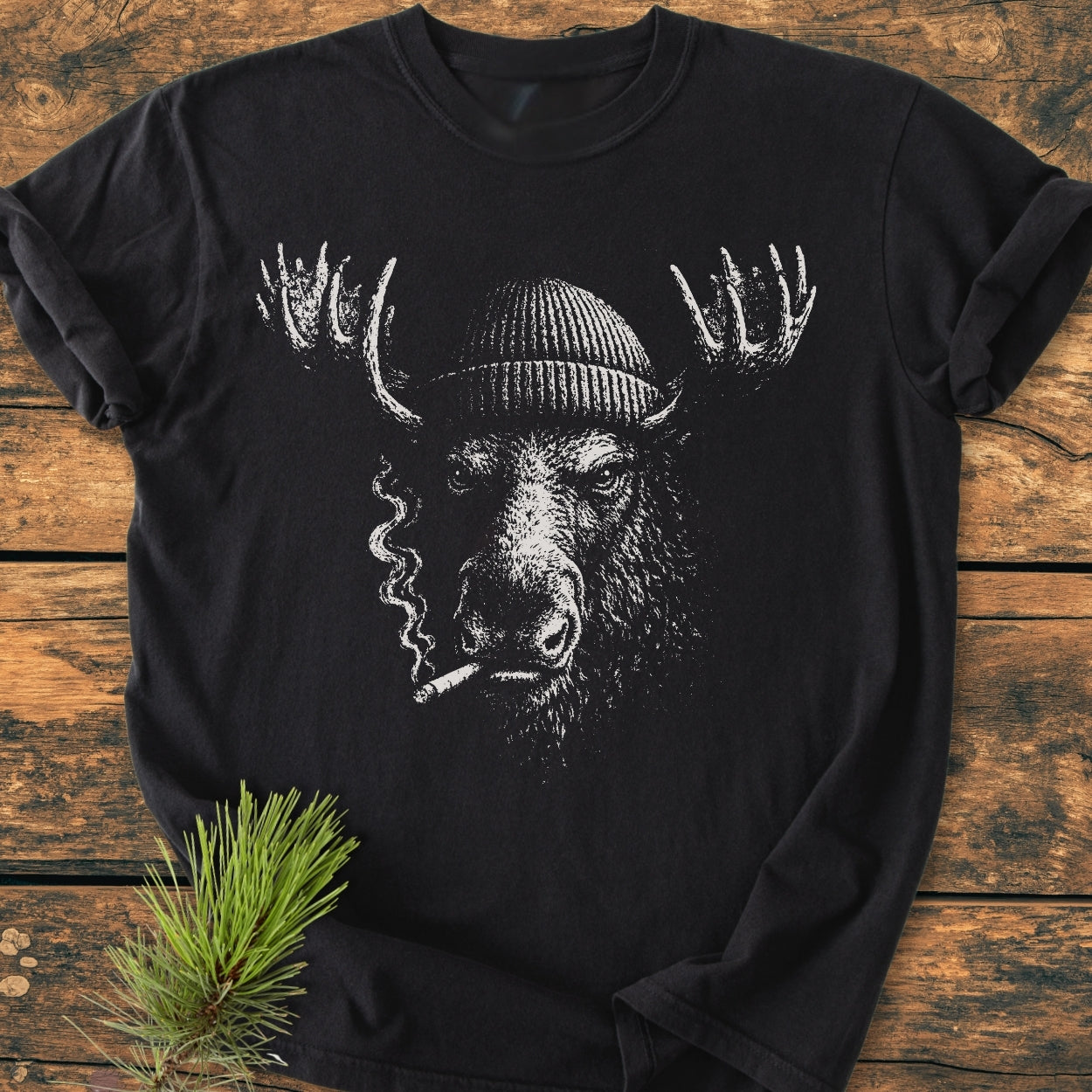 Slow Burn Moose - Vintage Wash Tee featuring a black T-shirt with a stylized moose head in a beanie, smoking, printed in white, laid on wood with a pine sprig nearby.