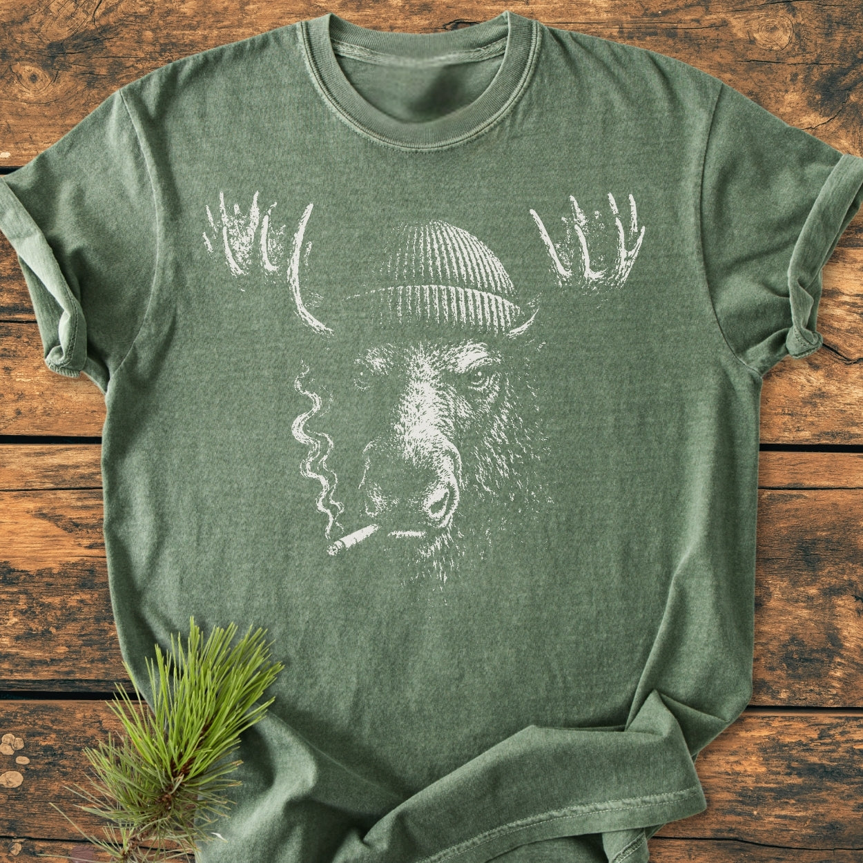 Slow Burn Moose - Vintage Wash Tee featuring a stylized moose with a knit cap and cigarette, screen-printed on a heather-green shirt, laid flat on rustic wood with pine sprig.