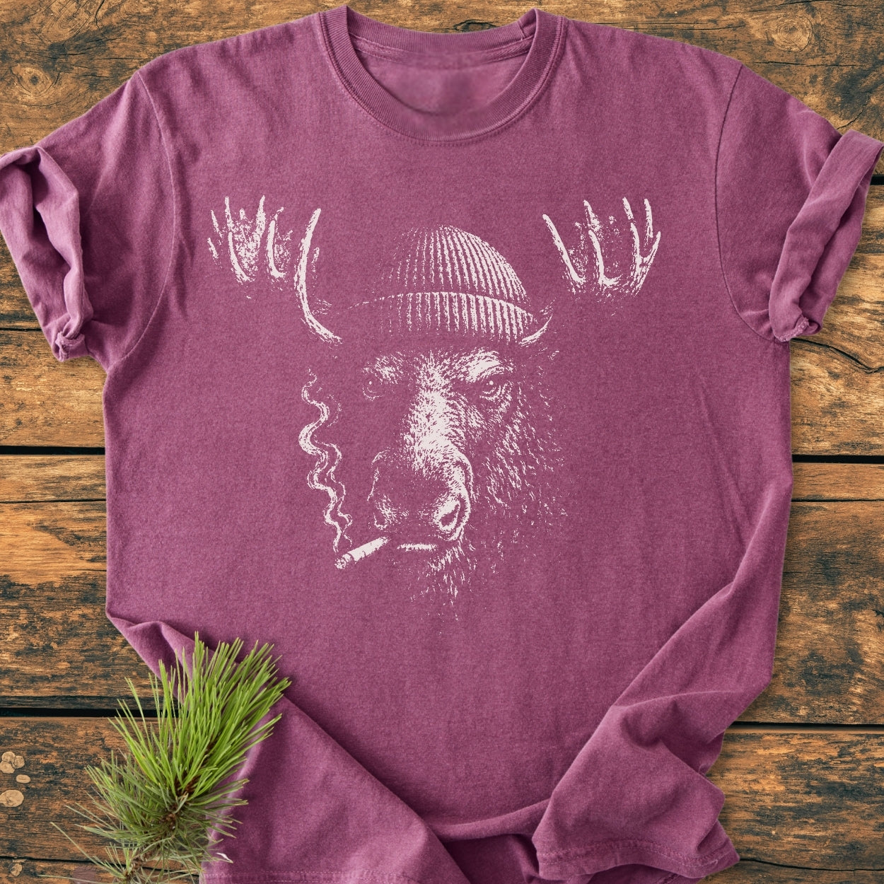 Slow Burn Moose - Vintage Wash Tee features a moose in a knit cap, smoking, on a heather-purple shirt, laid on wood, with a pine sprig accent.