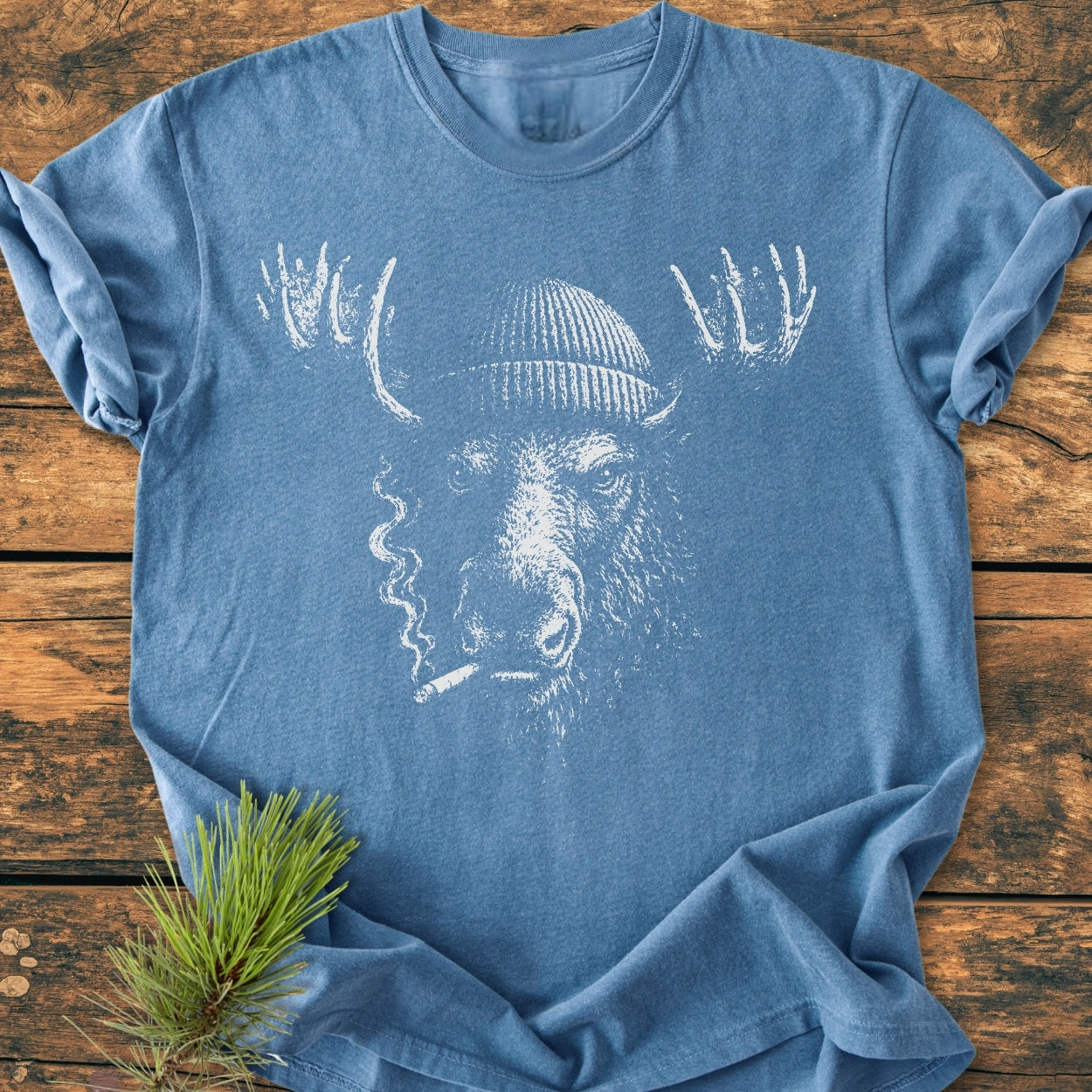 Slow Burn Moose - Vintage Wash Tee features a heather-blue hue with a white line drawing of a moose in a knit cap, cigarette in mouth, laid flat on a wooden surface.
