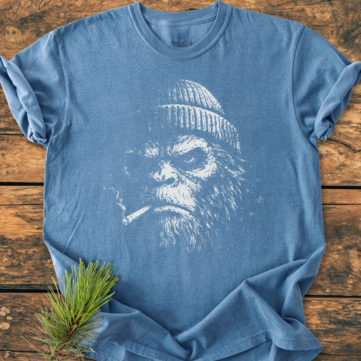 The Slow Burn Club - Vintage Wash Tee features a bearded creature print on a blue T-shirt, laid on a wooden surface with a pine needle sprig, highlighting its woodland-inspired design.