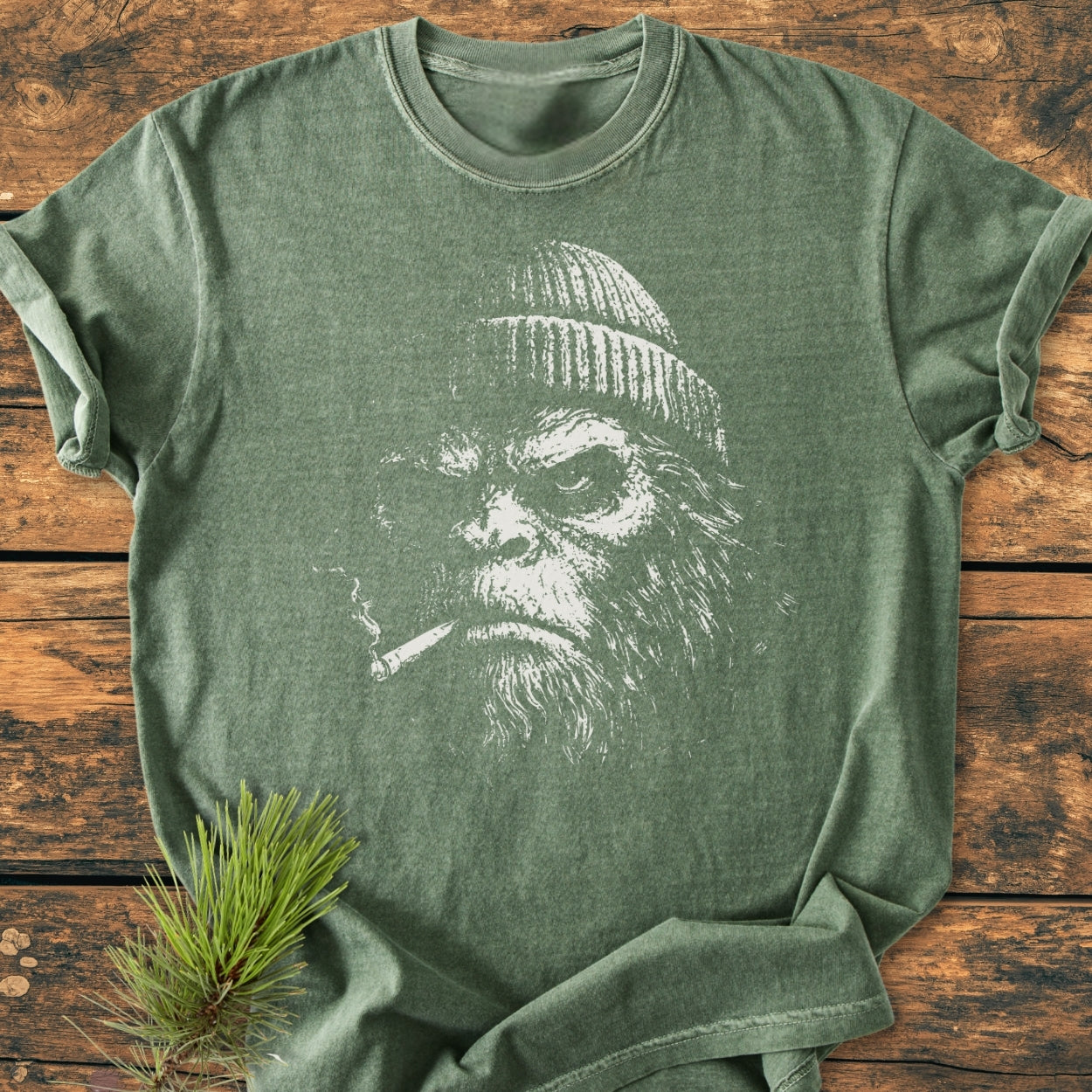 Slow Burn Club - Vintage Wash Tee featuring a bearded Bigfoot design in white, smoking a cigarette; laid flat on a wooden surface with a pine bough beside it.