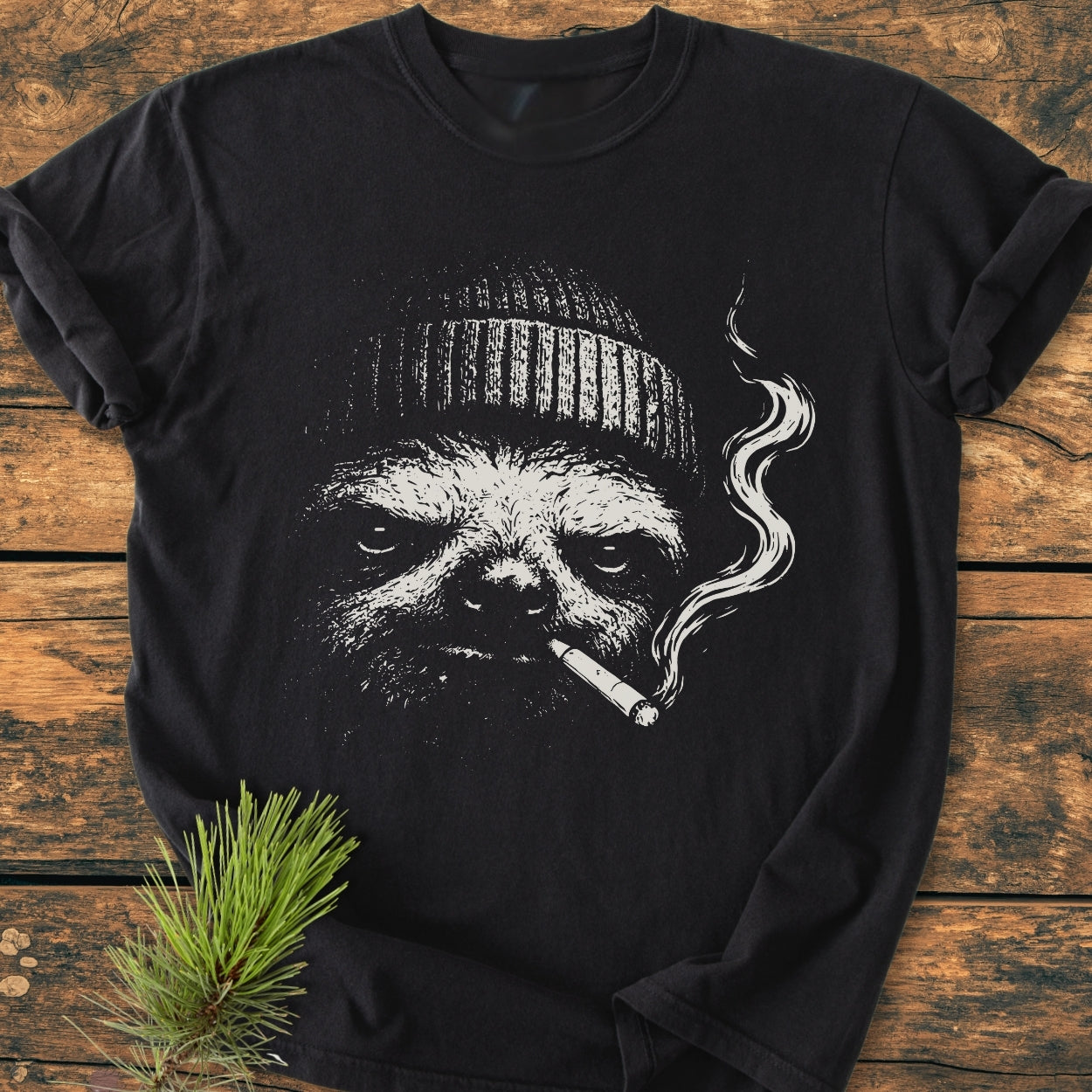 No Rush Club - Vintage Wash Tee featuring a sloth illustration with a beanie and cigarette, laid flat on wood. Includes pine needle accent. Made from soft, durable, garment-dyed cotton for comfort.