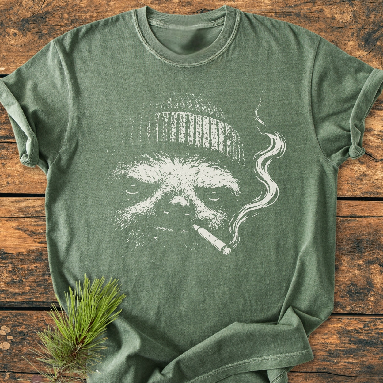 No Rush Club - Vintage Wash Tee features a white sketch of a sloth with a beanie and cigarette on a heather-green T-shirt, styled with a pine sprig on wooden surface.
