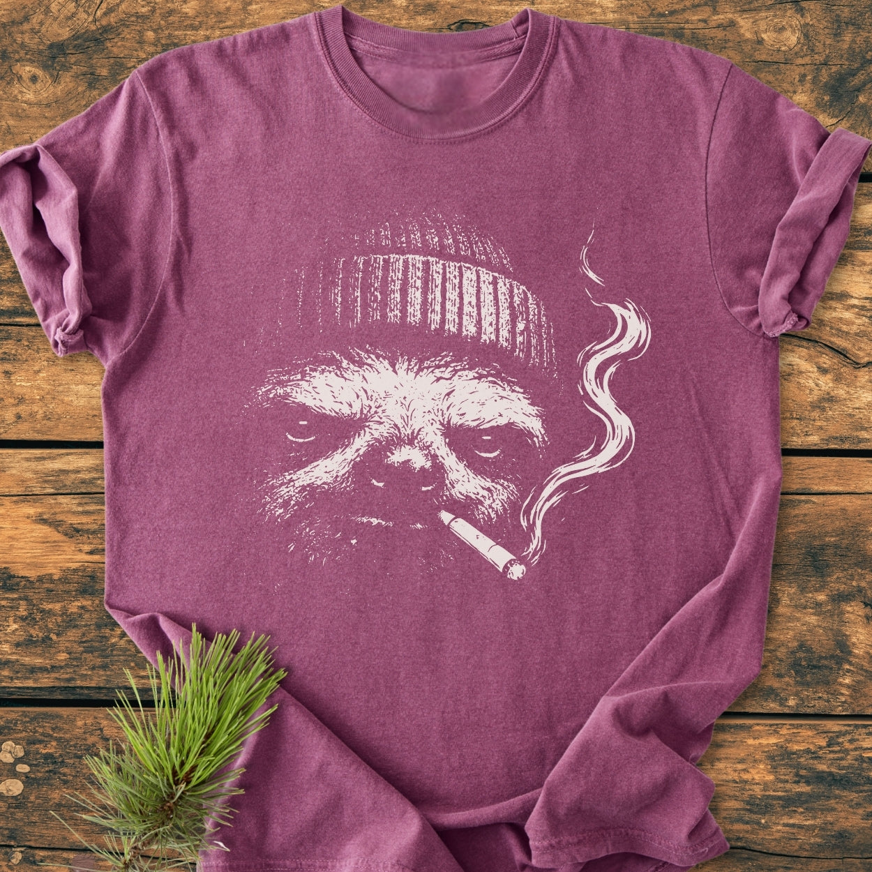 No Rush Club - Vintage Wash Tee features a sloth face with a beanie and cigarette, on a heathered T-shirt, displayed flat on a wooden surface with pine sprig accent.