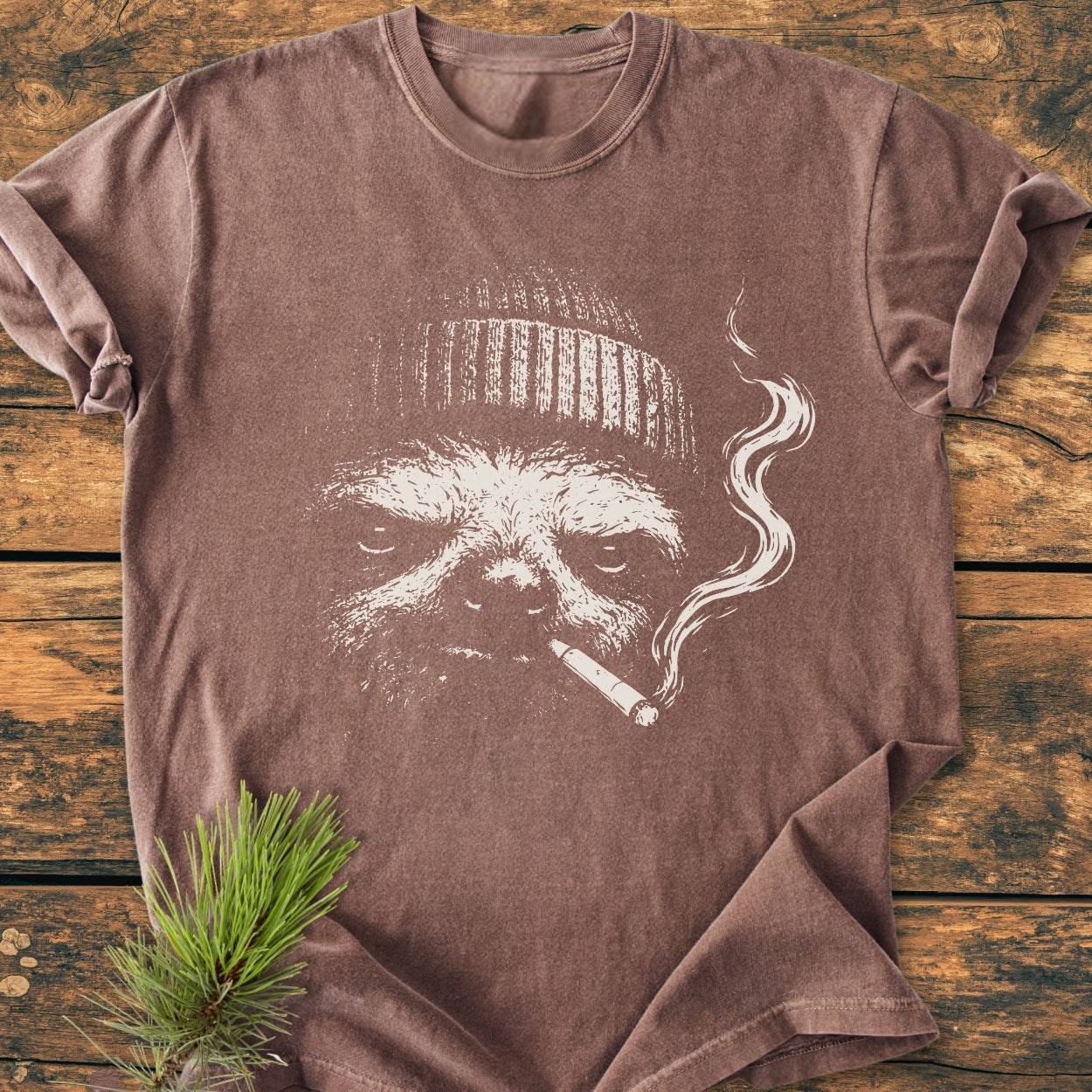 No Rush Club - Vintage Wash Tee featuring a stylized sloth face with a beanie and cigarette. Laid on wood, with pine sprig accent, highlighting the tee's relaxed fit and durable design.