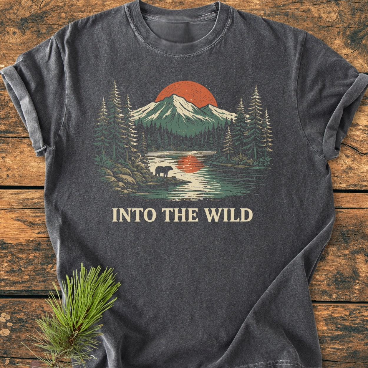 Into The Wild - Vintage Wash Tee featuring a stylized wilderness scene with mountains, a red sun, pine trees, a bear by a lake, and the caption. Laid on a wooden surface.