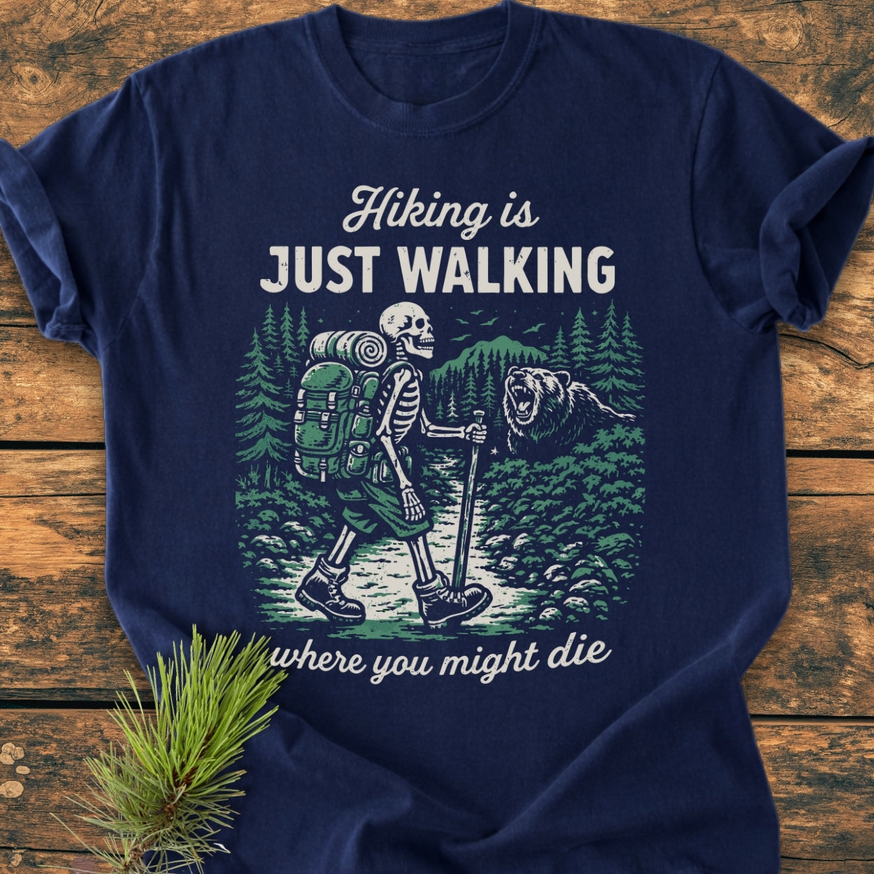 Navy-blue Hiking Is Just Walking - Vintage Wash Tee with a skeleton graphic hiking through a forest, featuring pine trees and a bear. Text reads: “Hiking is JUST WALKING where you might die.”
